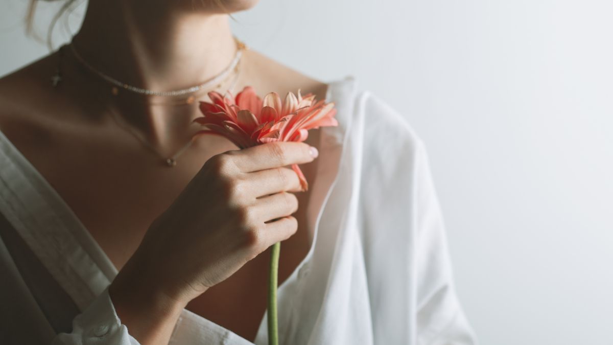Una donna tiene un fiore rosa vicino al viso, indossando una camicia bianca.
